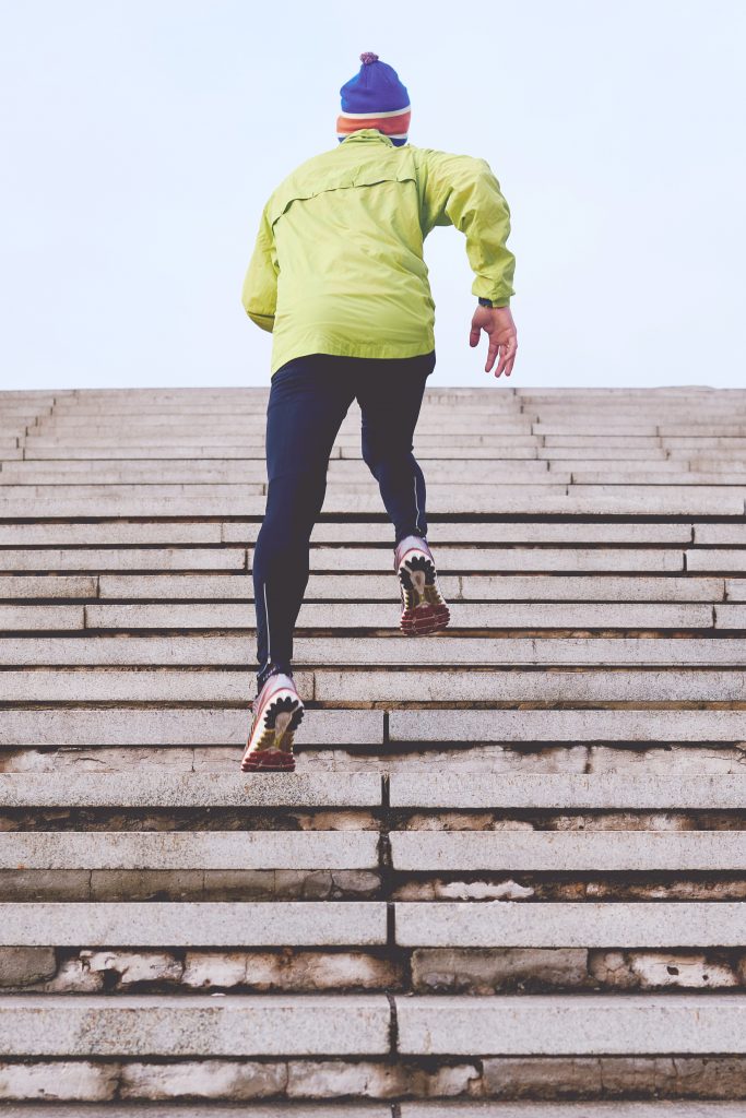 personne de dos faisant du running dans des escaliers portant un bonnet une veste verte un collant de sport et des baskets